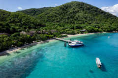 A boat docked at a pier near a tropical resort with lush green hills and clear turquoise water.