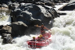 A group of people in a red raft navigating through rough whitewater rapids with a guide standing at the back cheering.