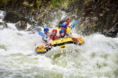 Group of rafters in helmets paddle a yellow raft through crashing whitewater rapids, water splashing around them.